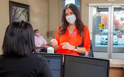 Woman with an orange shirt and face mask talking to a woman with a black shirt at the reception desk while socially distanced.